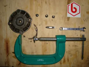 C-clamp, mata obeng, kunci, sekrup, dan penutup logam di atas papan.