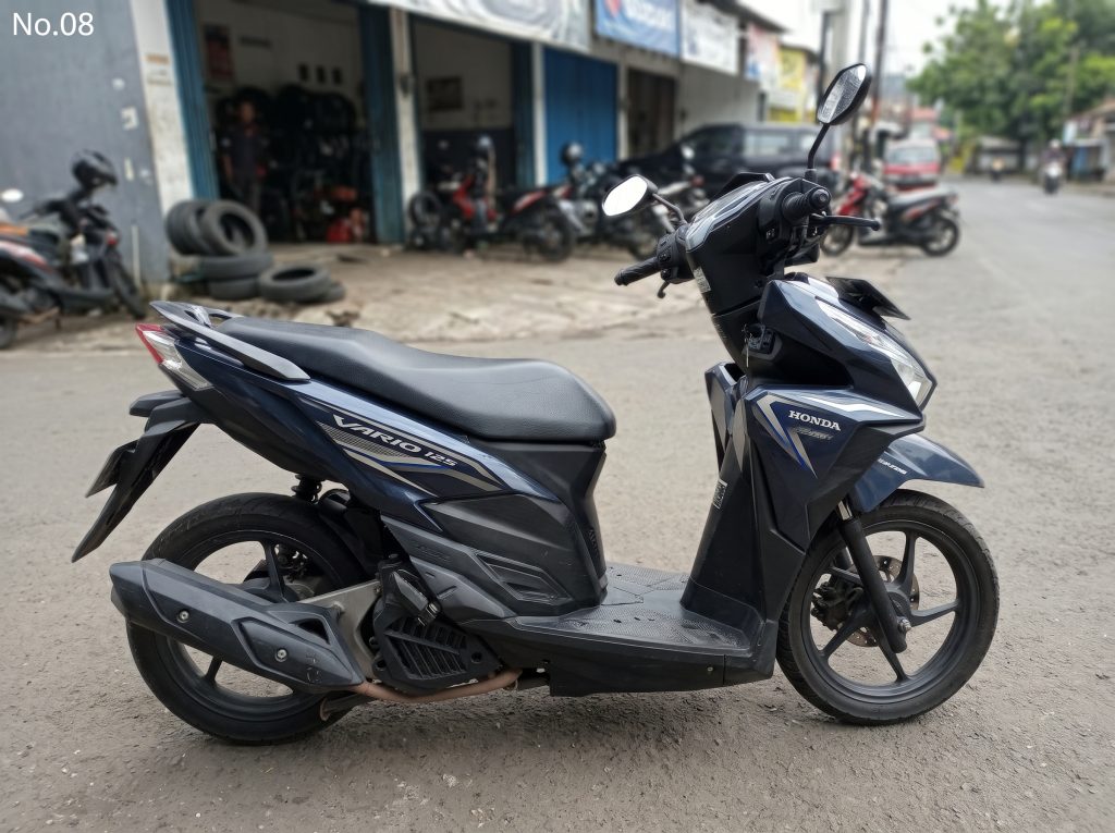 Skuter biru gelap bertuliskan Vario 125 yang diparkir di depan deretan bengkel.